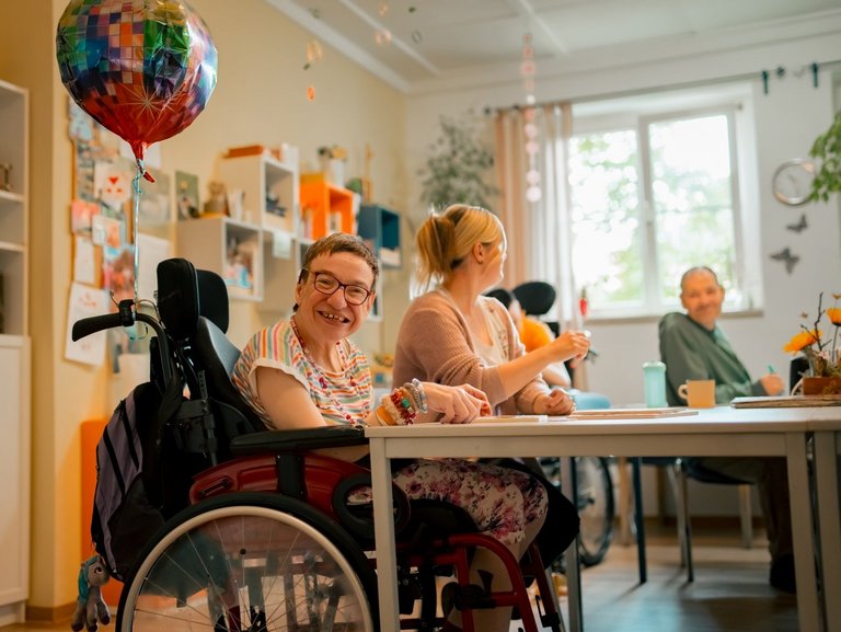 Eine Gruppe von Menschen mit und ohne Beeinträchtigung sitzen am Tisch und unterhalten sich miteinander, während sie gemeinsam Spiele spielen oder an kreativen Projekten arbeiten.