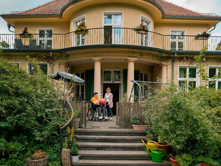 Ein junger Mensch im Rollstuhl und eine Frau unterhalten sich auf der Terrasse eines gelben Hauses mit Balkon. Im Vordergrund sind Pflanzen und Gießkannen in einem Garten zu sehen.
