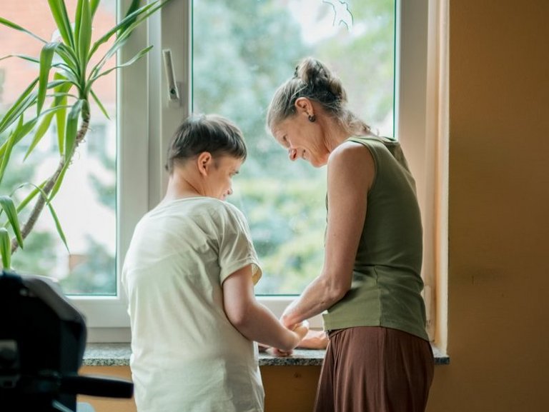 Eine Person mit Behinderung und eine Mitarbeitende stehen gemeinsam am Fenster und unterhalten sich lachend.