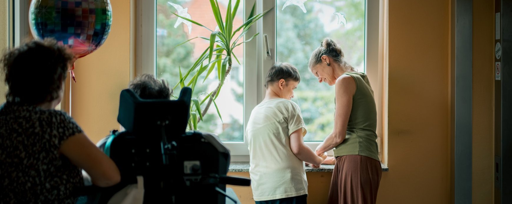 Eine Person mit Behinderung und eine Mitarbeitende stehen gemeinsam am Fenster und unterhalten sich lachend.