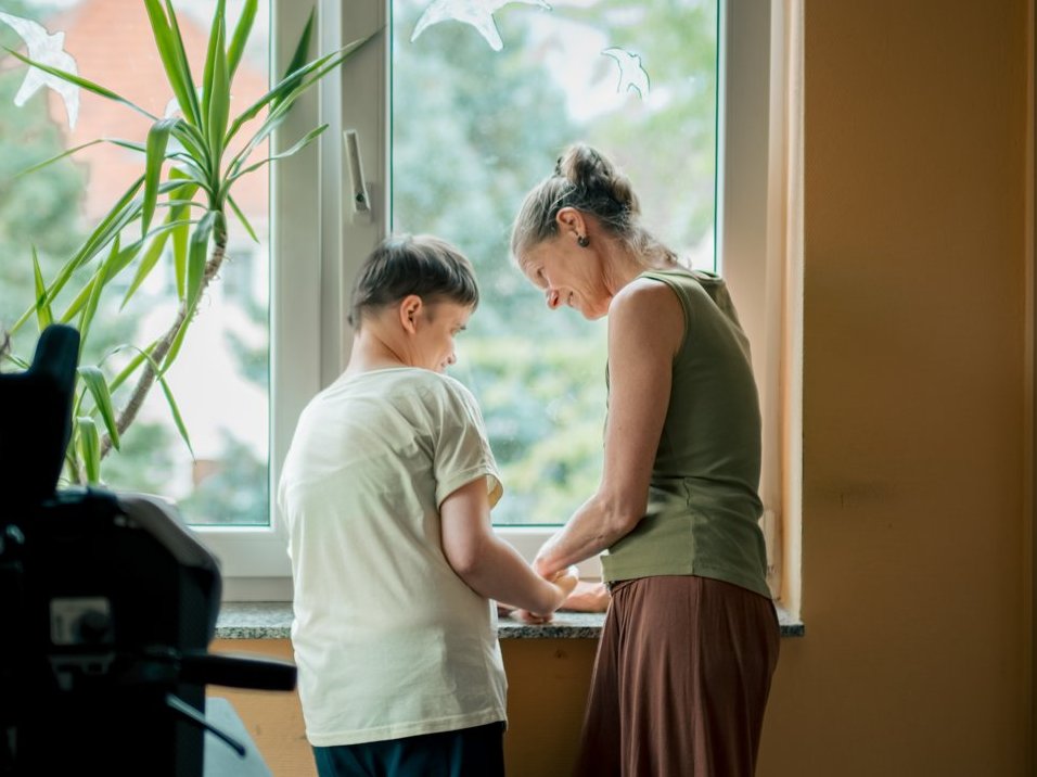 Eine Person mit Behinderung und eine Mitarbeitende stehen gemeinsam am Fenster und unterhalten sich lachend.