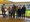 Sieben Personen posieren für ein Gruppenfoto auf einer Tribüne in einem Fußballstadion. Im Hintergrund sind gelbe Sitzreihen und der grüne Rasen zu erkennen.