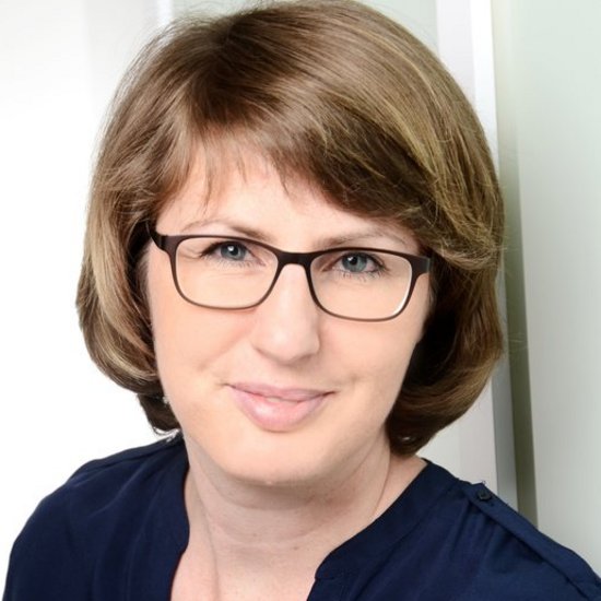 Porträt einer lächelnden Frau mit kurzen braunen Haaren und Brille. Sie trägt ein dunkelblaues Oberteil und blickt direkt in die Kamera.