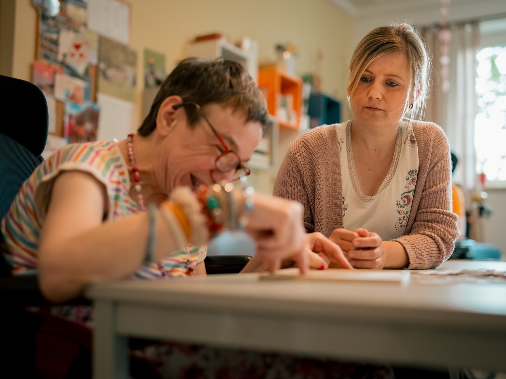 Eine Bewohnerin mit Beeinträchtigung sitzt mit einer Mitarbeiterin am Tisch und setzt schmunzelnd ein Puzzle zusammen. 