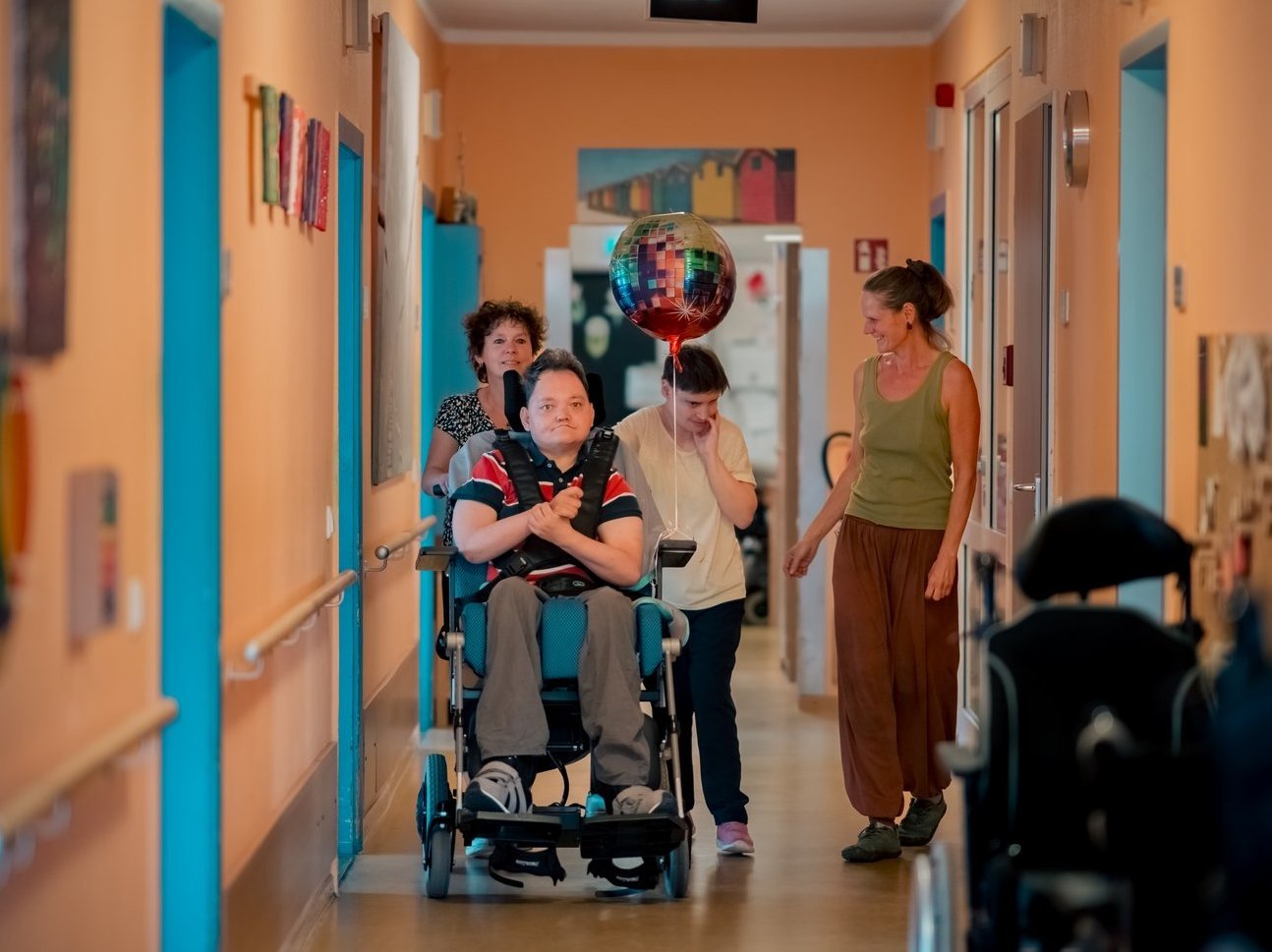 Mitarbeitende und Bewohnende, darunter Personen im Rollstuhl, gehen bzw. fahren über den Flur und unterhalten sich.