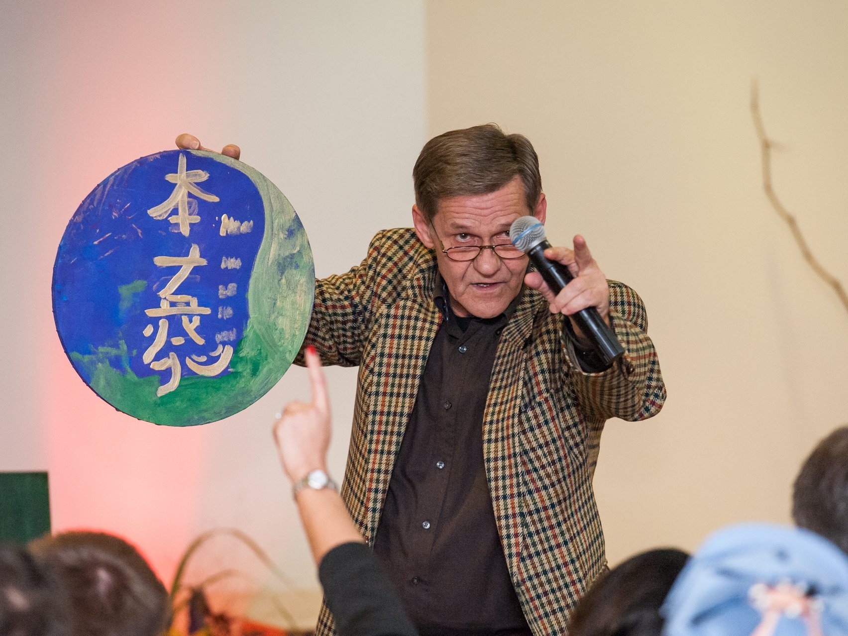 Ein älterer Mann mit karriertem Sakko und Mikrofon spricht zum Publikum und hält dabei eine runde Tafel mit chinesischen Schriftzeichen in die Luft