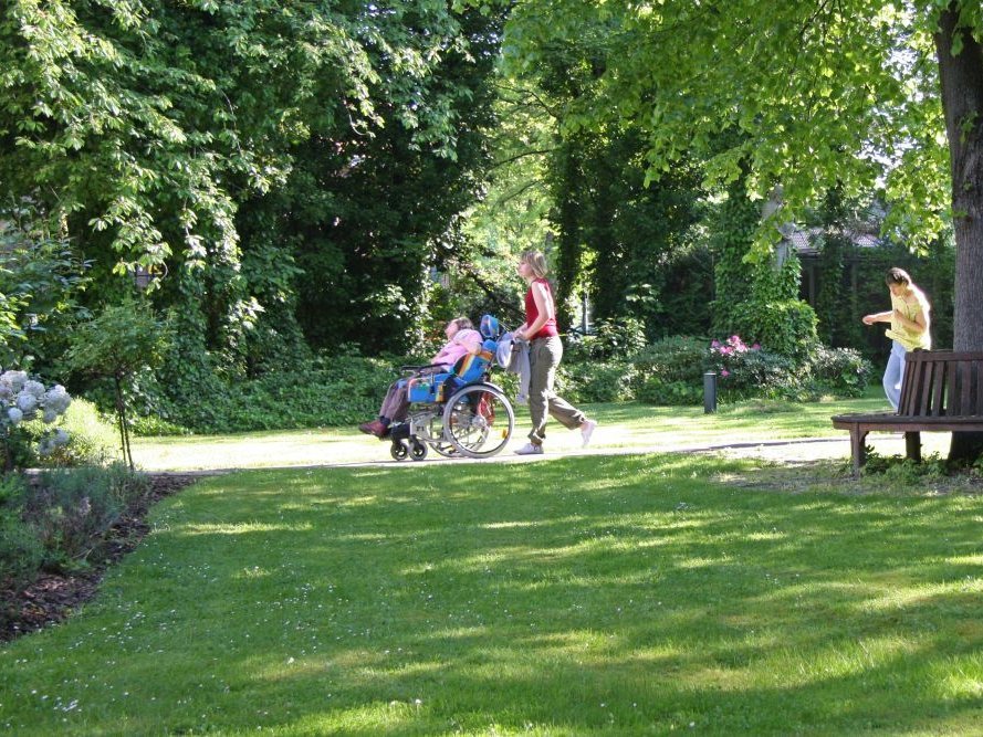 Spazieren im Park: Eine Frau schiebt einen Rollstul mit einer Bewohnerin
