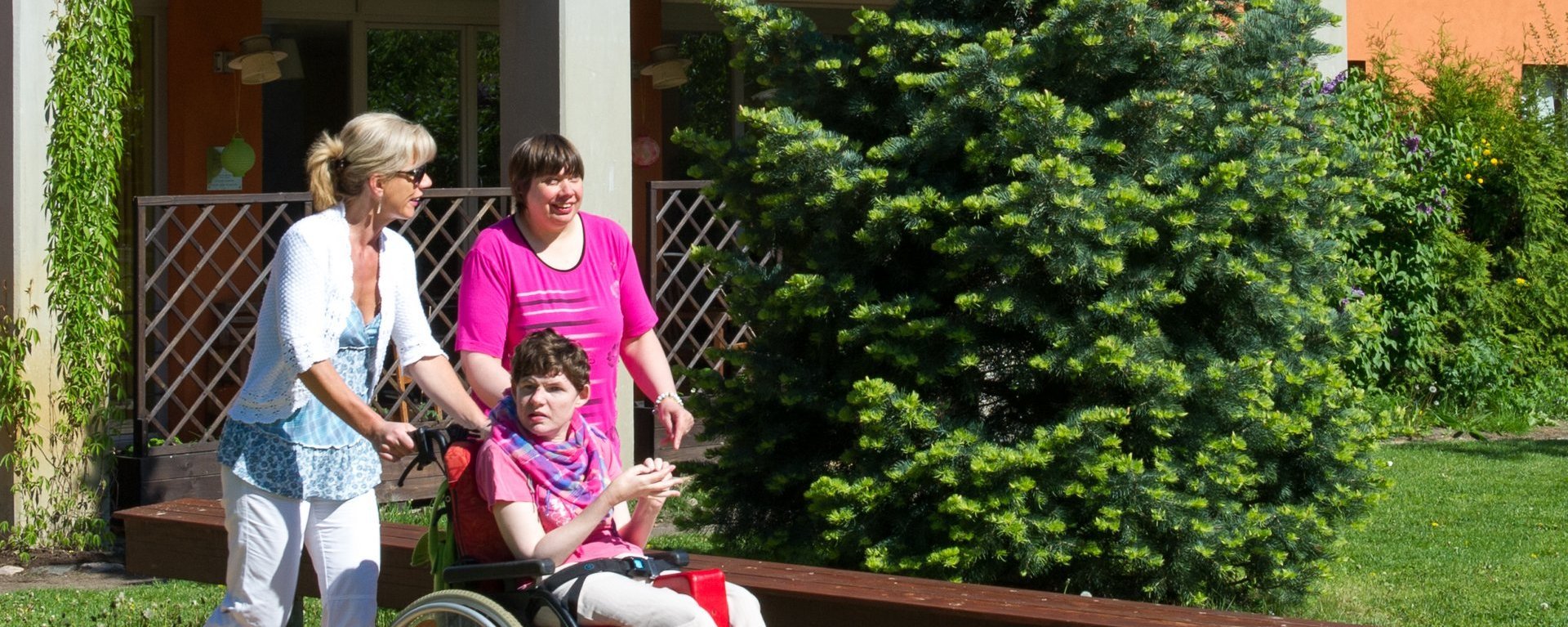 schönes Wetter: eine Betreuerin schiebt eine Frau im Rollstuhl und unterält sich mit einer weiteren Frau