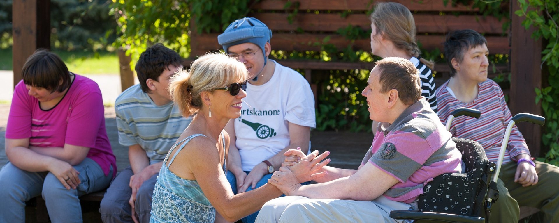 Menschen mit und ohne geistige und körperliche Beeinträchtigung sitzen im Garten im Schatten und unterhalten sich.