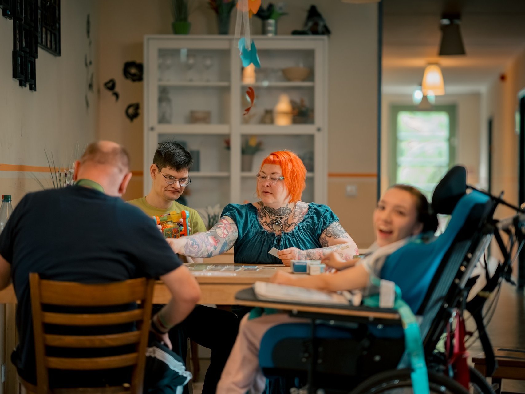 Eine Gruppe von Menschen mit und ohne Beeinträchtigungen sitzt am Tisch. Einige lachen, andere sind vertieft in ihre Beschäftigung.