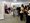 Eine Gruppe von Menschen besucht eine Ausstellung. Sie stehen vor weißen Präsentationswänden, auf denen Fotos und Texte zu sehen sind. Ein großes Foto zeigt eine Frau und einen Mann, die auf unterschiedlich hohen Stühlen sitzen. Die Besucher wirken interessiert.